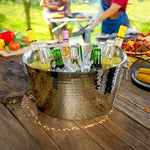 Round large ice bucket chilling beer and wine on a patio table at a barbecue.  There are people grilling in the background.